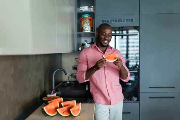 Multiracial man eating watermelon in his kitchen during hot summer.