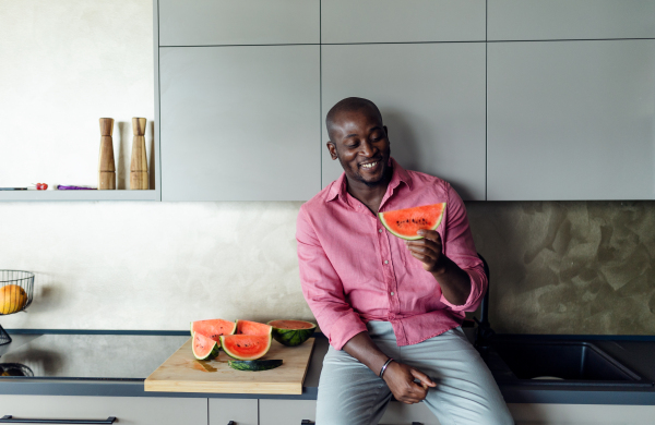 Multiracial man eating watermelon in his kitchen during hot summer.