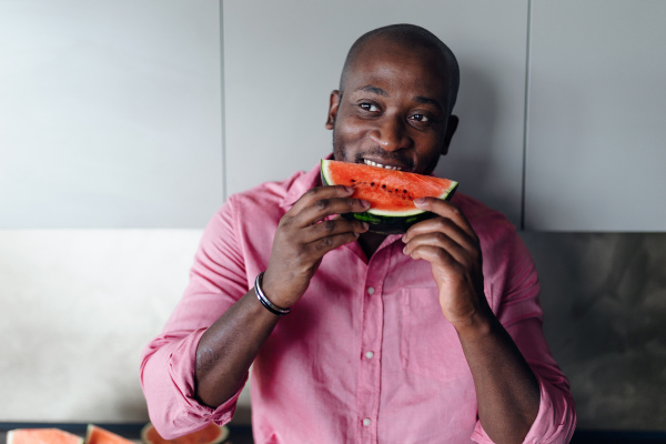 Multiracial man eating watermelon in his kitchen during hot summer.