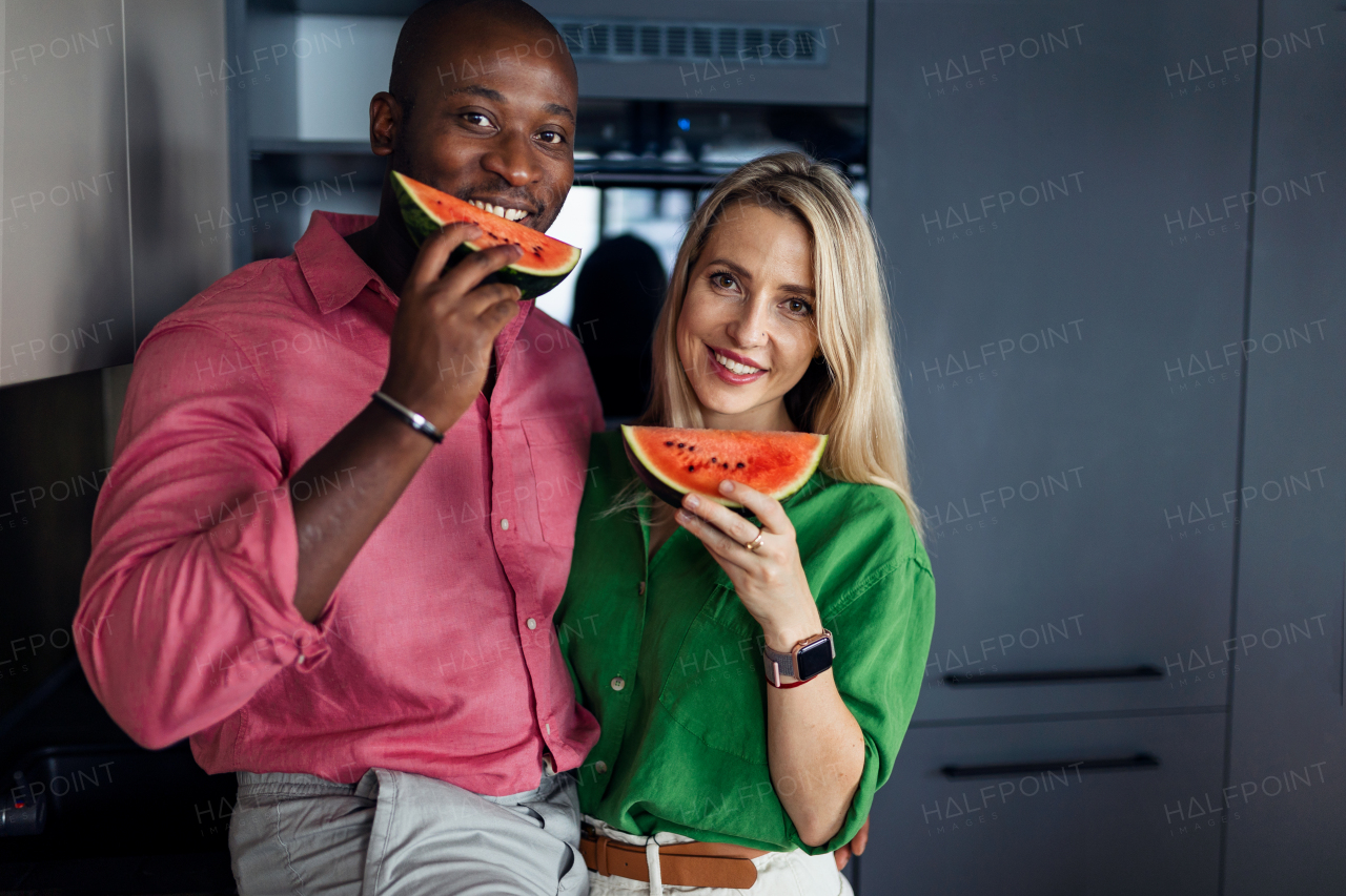 Multiracial couple eating a melon in kitchen during hot sunny days.