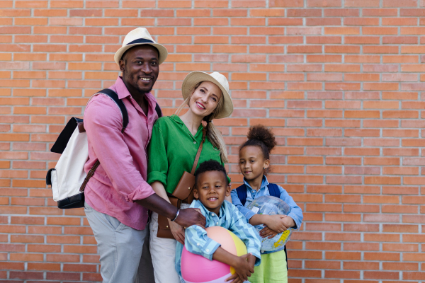 Multiracial family travelling together with small kids. Posing in front of a brick wall with beach ball.