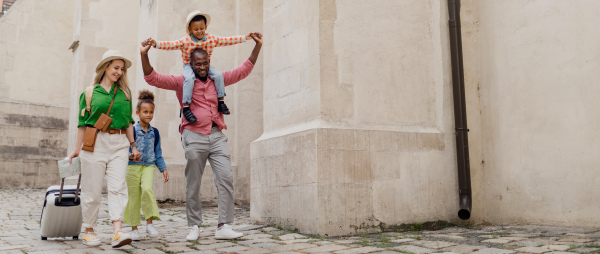 Happy multiracial family travel together with suitcases, walking in old city centre.