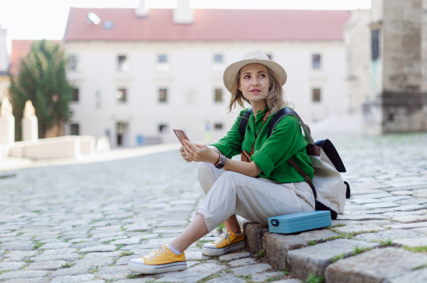 Young blond woman travel alone in old city centre, sitting and using smartphone.