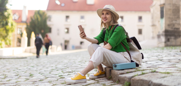 Young blond woman travel alone in old city centre, sitting and using smartphone.