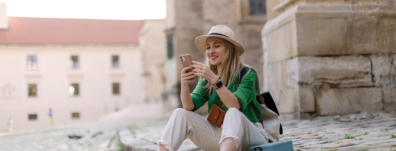 Young blond woman travel alone in old city centre, sitting and using smartphone.