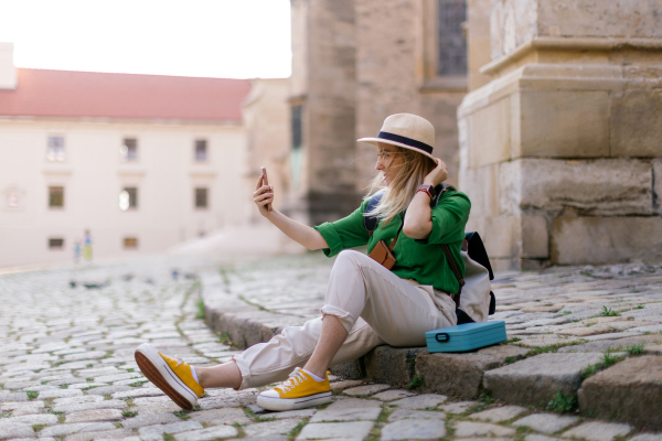 Young blond woman travel alone in old city centre, sitting and using smartphone, taking selfie.