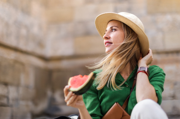 A young woman traveller eating watermelon in street during hot sunny day, summer vacation trip concept.