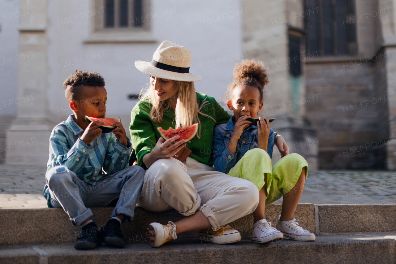 Multiracial kids with mother eating a watermelon in street during hot sunny days, summer holiday travelling concept.