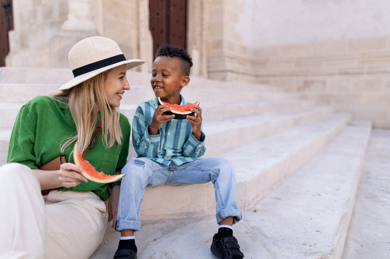 Multiracial son with mother eating a watermelon in street during hot sunny days, summer holiday travelling concept.