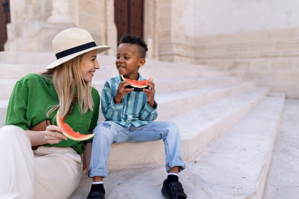 Multiracial son with mother eating a watermelon in street during hot sunny days, summer holiday travelling concept.