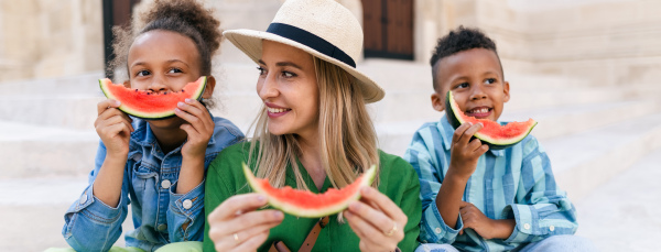 Multiracial kids with mother eating a watermelon in street during hot sunny days, summer holiday travelling concept.