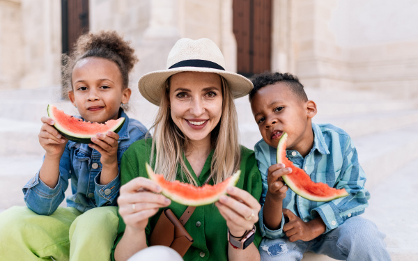 Multiracial kids with mother eating a watermelon in street during hot sunny days, summer holiday travelling concept.