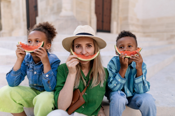 Multiracial kids with mother eating a watermelon in street during hot sunny days, summer holiday travelling concept.