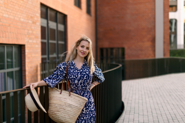 Young woman with straw bag in a city during sunset, standing and posing in front of old brick building.