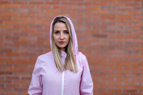 Young blond woman with hood on head, standing in front of a brick wall.