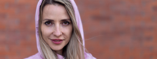 Wide photography of young blond woman with hood on head, standing in front of a brick wall.