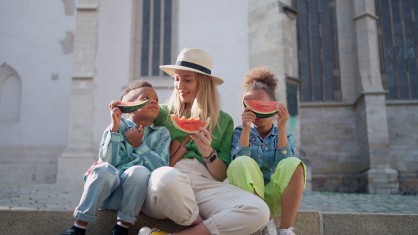 Multiracial kids with mother eating a watermelon in street during hot sunny days, summer holiday travelling concept.