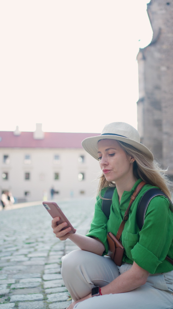 A young woman travel alone in old city centre, sitting and using smartphone, vertical footage.