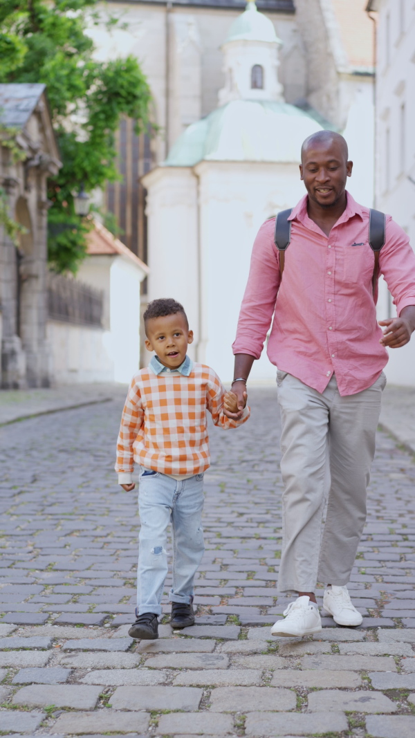 A multiracial father holding his little son and walking in town in summer together, vertical footage.