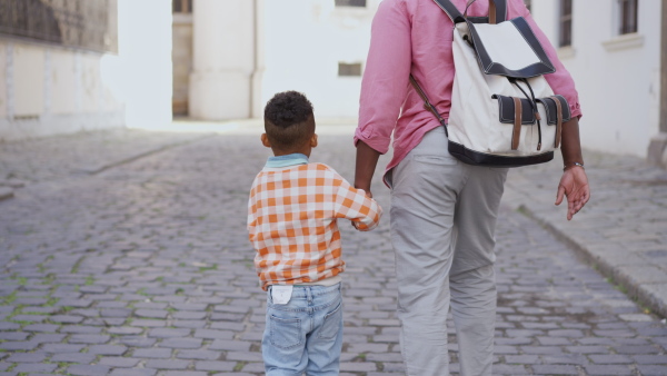 A multiracial father holding his little son and walking in town in summer together, rear view.