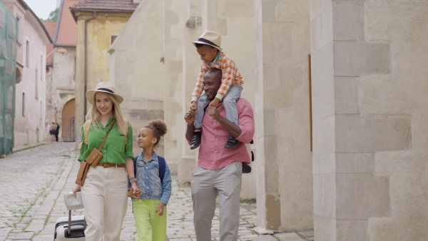 Happy multiracial family travel together with suitcases, walking in old city centre.