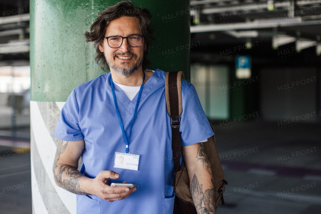 Male doctor in uniform going home from work, standing on parking lot, looking at camera. Work-life balance of healthcare worker.