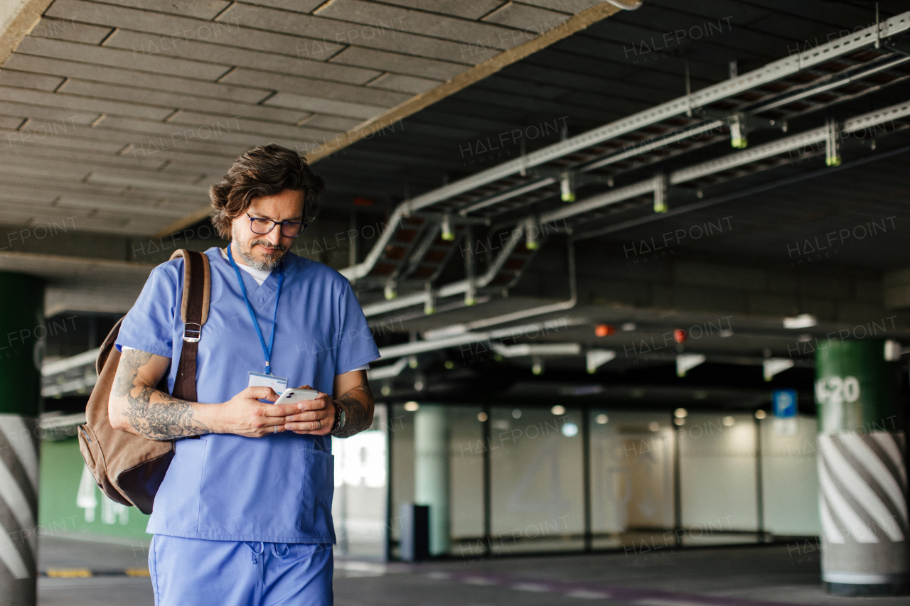 Male doctor in uniform in parking lot, going home from work, texting on smartphone. Work-life balance of healthcare worker.