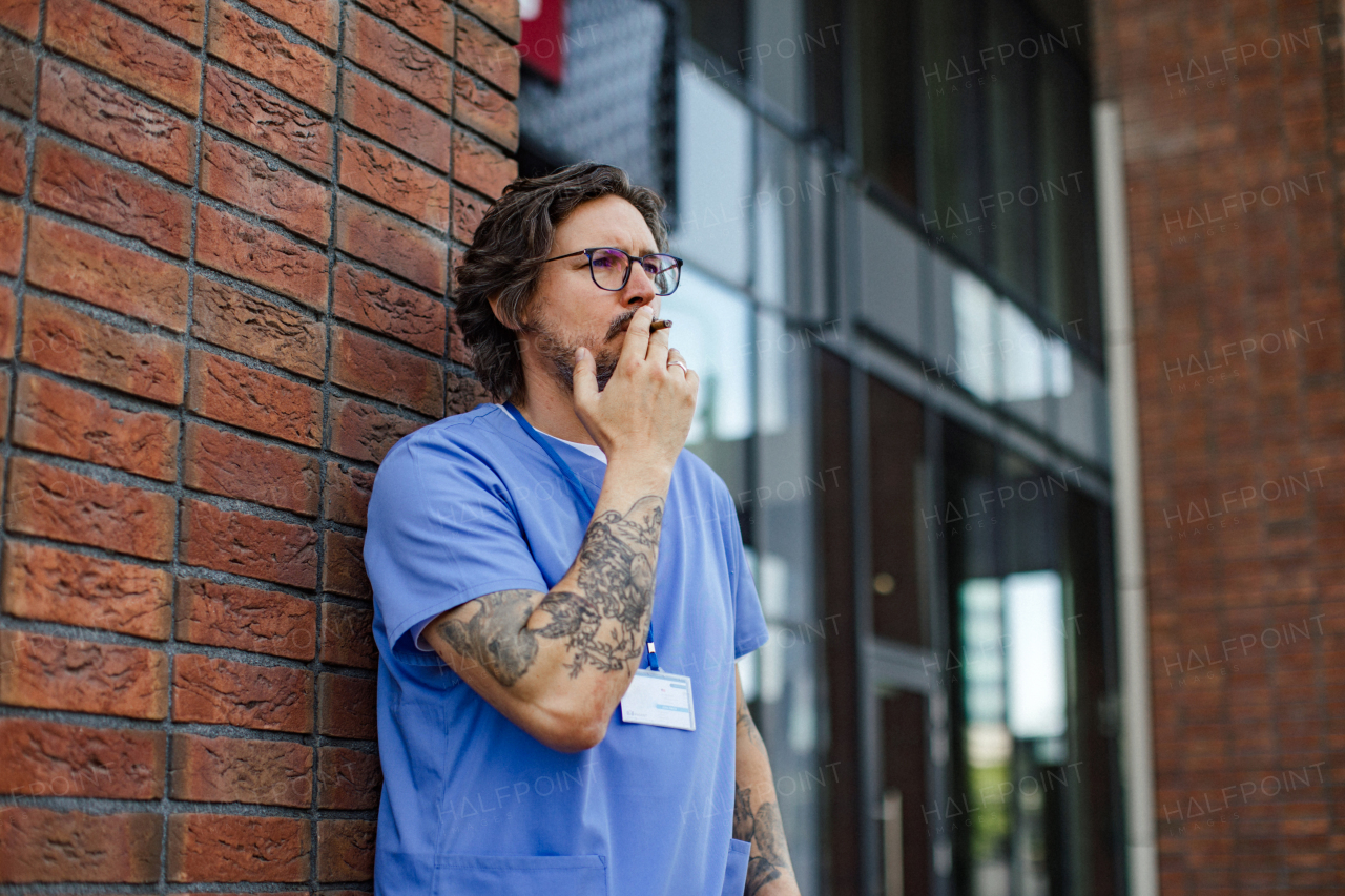 Healthcare worker taking a break, smoking a cigarette outdoors, in front of the hospital building. Handsome male doctor or nurse.