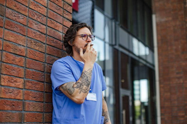 Healthcare worker taking a break, smoking a cigarette outdoors, in front of the hospital building. Handsome male doctor or nurse.