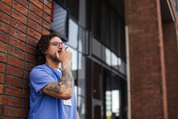 Healthcare worker taking a break, smoking a cigarette outdoors, in front of the hospital building. Handsome male doctor or nurse.