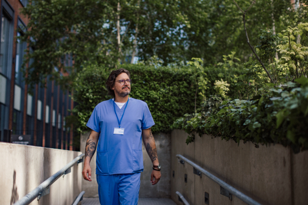 Male n uniform going home from work, walking in front of hospital building. Work-life balance of healthcare worker.