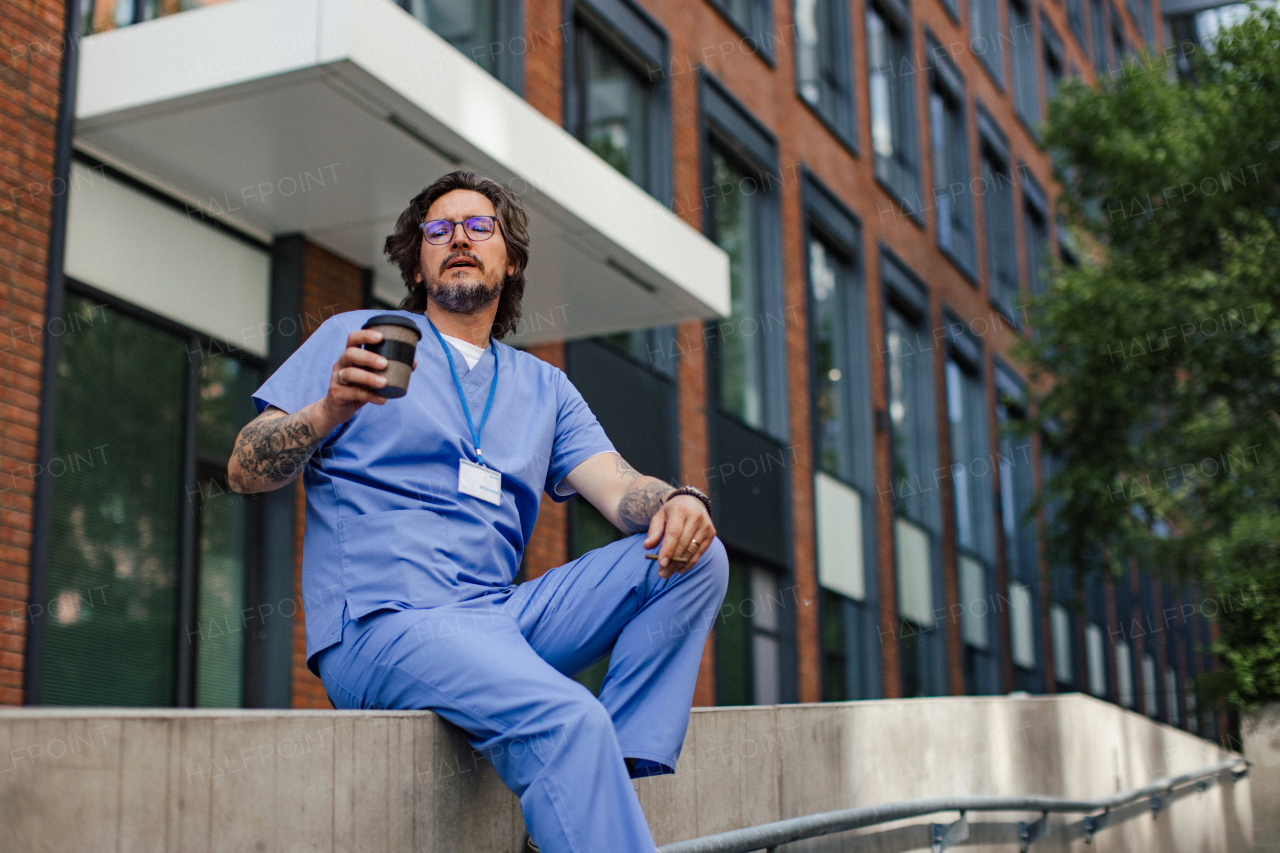 Healthcare worker taking a break, smoking a cigarette outdoors, drinking coffee from traveling mug, in front of the hospital building. Handsome male doctor or nurse.