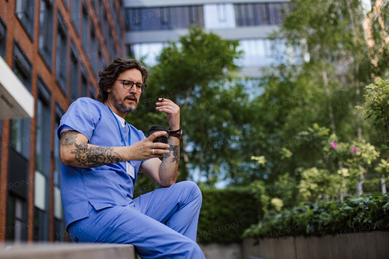 Healthcare worker taking a break, smoking a cigarette outdoors, scrolling on phone, in front of the hospital building. Handsome male doctor or nurse.