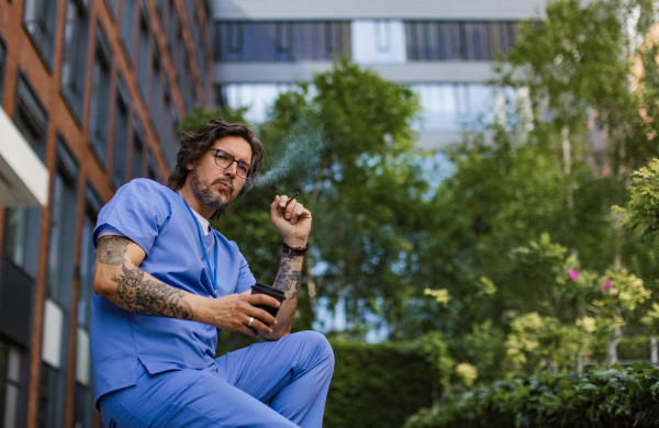 Healthcare worker taking a break, smoking a cigarette outdoors, drinking coffee from traveling mug, in front of the hospital building. Handsome male doctor or nurse.