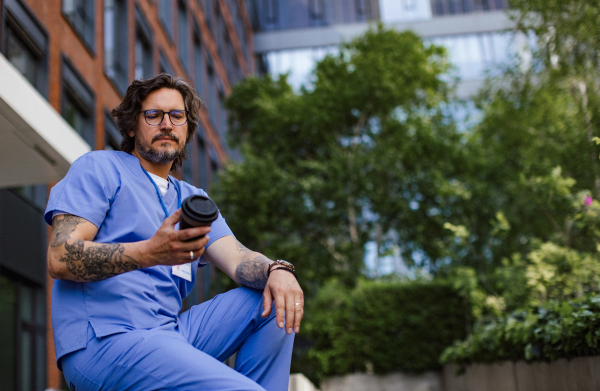 Healthcare worker taking a break, sitting outdoors, drinking coffee from traveling mug, in front of the hospital building. Handsome male doctor or nurse.