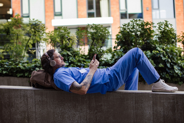 Doctor lying on low concrete wall, listening music, wearing headphones, scrolling on smartphone. Winding down from busy workday in hospital. Mental health and life balance for a healthcare worker.