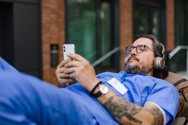 Doctor lying on park bench, listening music, wearing headphones, scrolling on smartphone. Winding down from busy workday in hospital. Mental health and life balance for a healthcare worker.