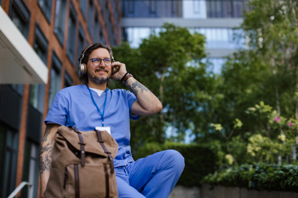 Doctor sitting on low concrete wall, listening music, wearing headphones. Winding down from busy workday in hospital. Mental health and life balance for a healthcare worker.