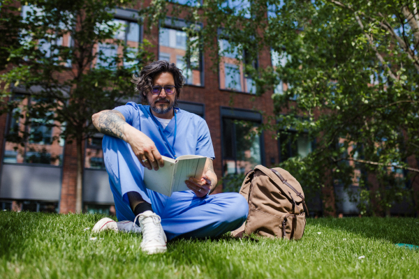 Handsome doctor sitting on grass, reading book and relaxing, winding down from busy workday in hospital. Mental health and life balance for a healthcare worker.