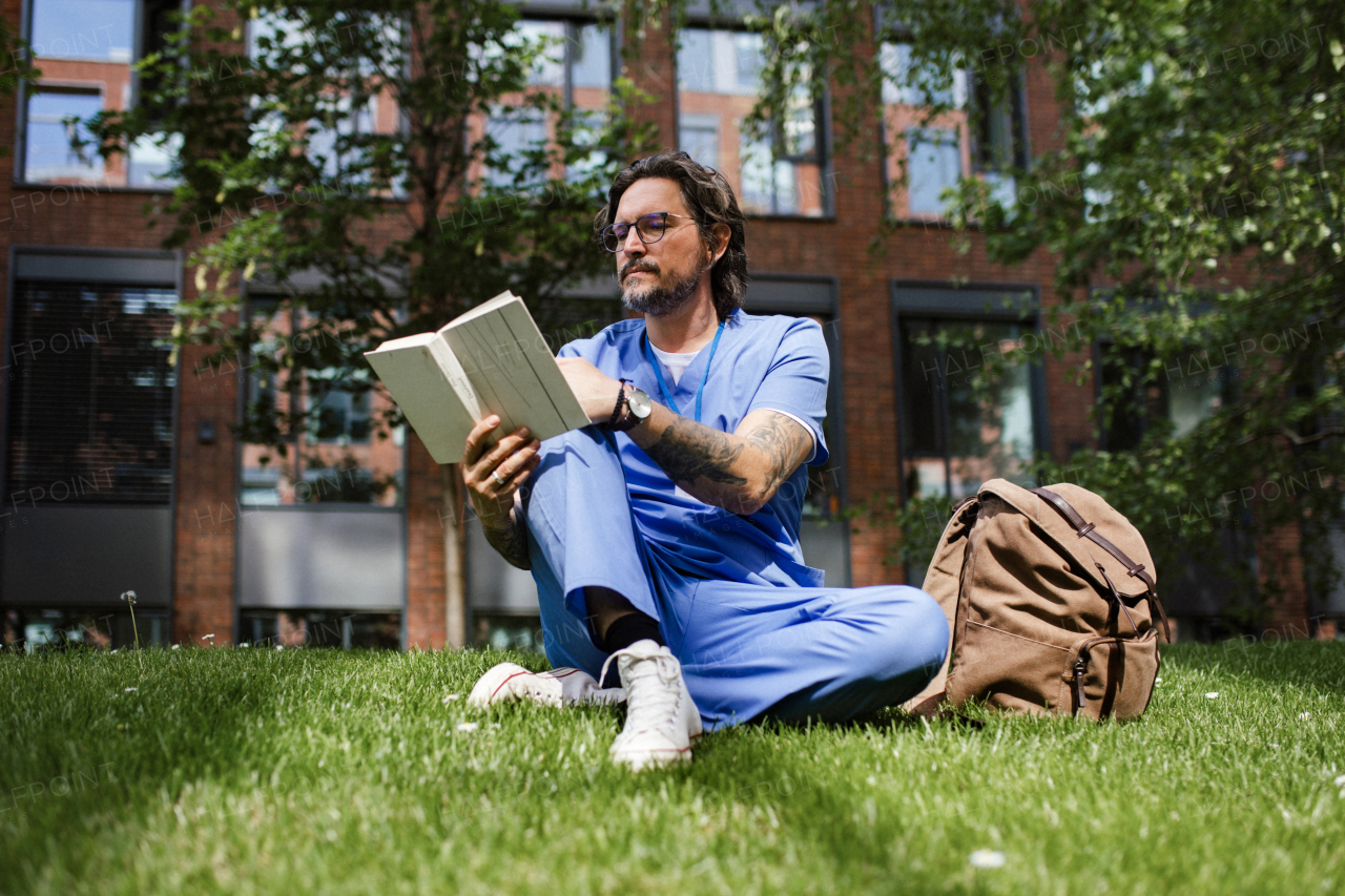 Handsome doctor sitting on grass, reading book and relaxing, winding down from busy workday in hospital. Mental health and life balance for a healthcare worker.