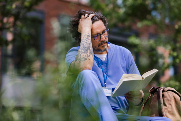 Handsome doctor sitting on grass, reading book and relaxing, winding down from busy workday in hospital. Mental health and life balance for a healthcare worker.