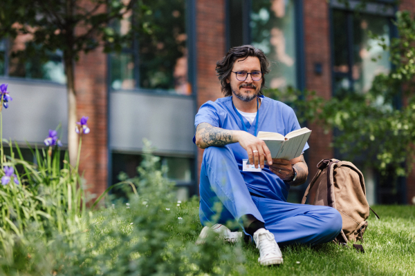 Handsome doctor sitting on grass, reading book and relaxing, winding down from busy workday in hospital. Mental health and life balance for a healthcare worker.