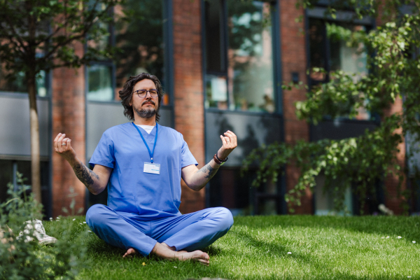 Handsome doctor sitting on grass, meditating and relaxing, winding down from busy workday in hospital. Mental health and life balance for a healthcare worker.