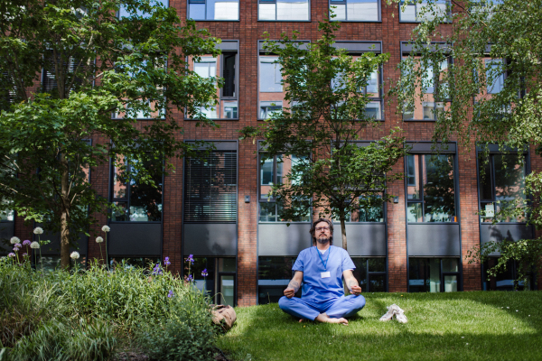 Handsome doctor sitting on grass, meditating and relaxing, winding down from busy workday in hospital. Mental health and life balance for a healthcare worker.