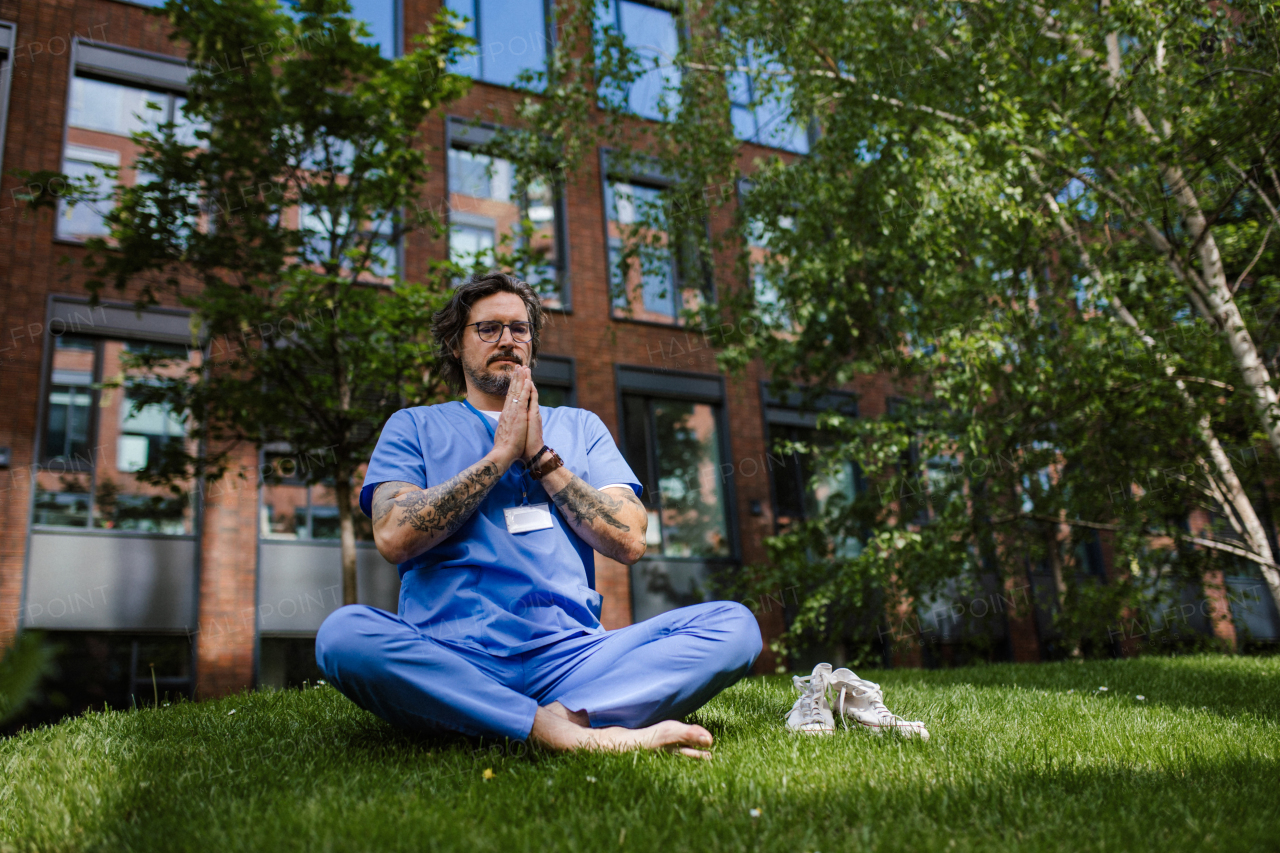 Handsome doctor sitting on grass, meditating and relaxing, winding down from busy workday in hospital. Mental health and life balance for a healthcare worker.