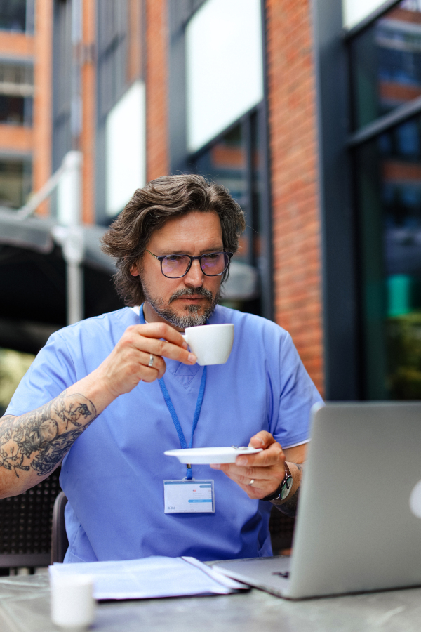 Doctor drinking coffee in coffee shop, working on laptop. Mental health and life balance for a healthcare worker. Telemedicine consultation online.