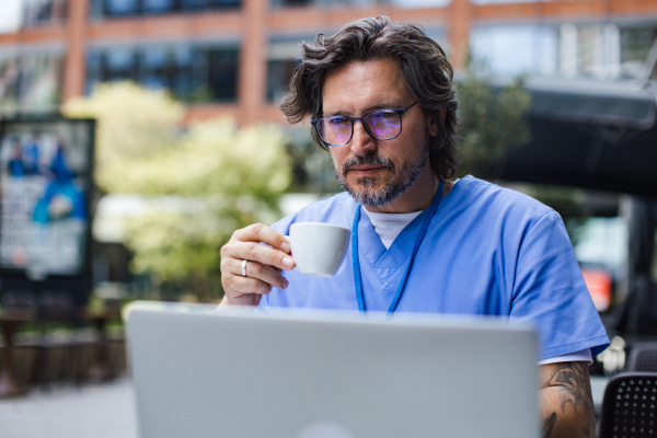 Doctor drinking coffee in coffee shop, working on laptop. Mental health and life balance for a healthcare worker. Telemedicine consultation online.