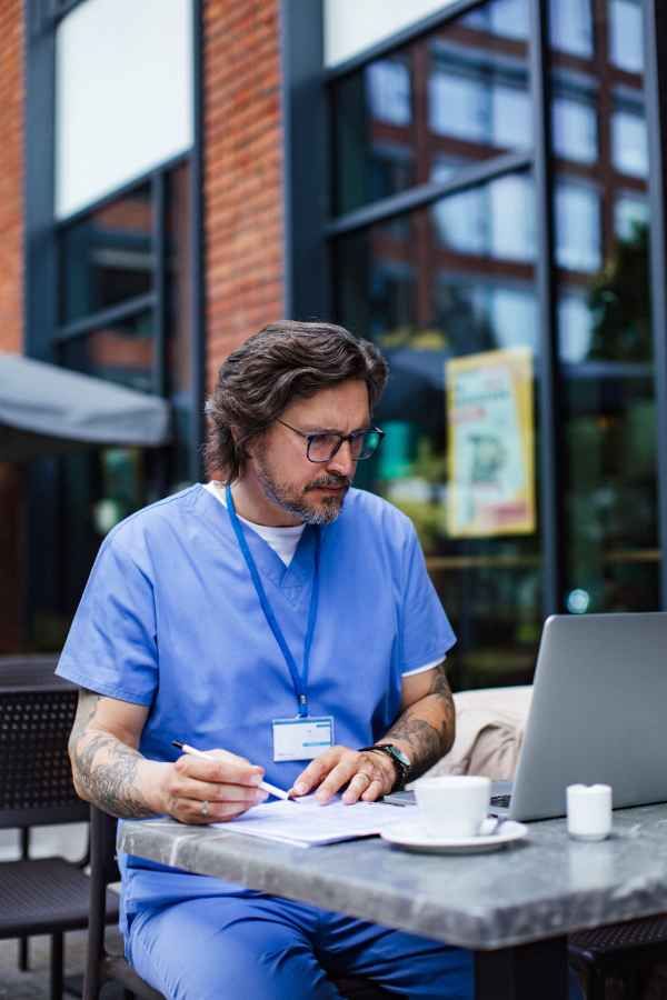 Doctor drinking coffee in coffee shop, working on laptop. Mental health and life balance for a healthcare worker. Telemedicine consultation online.