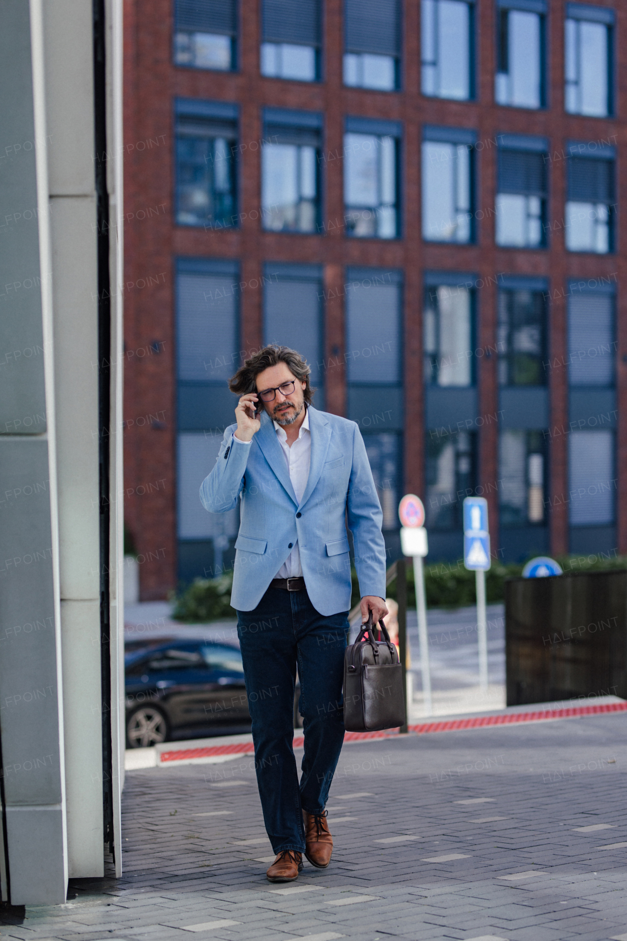 Portrait of mature businessman phone calling, standing on city street, in front of office building. Handsome man with glasses in a suit.