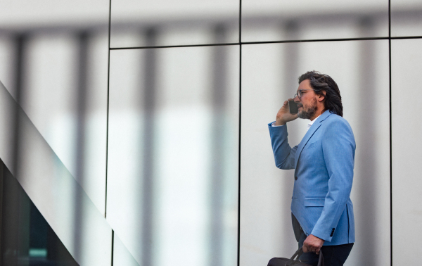 Portrait of mature businessman phone calling, standing on city street, in front of office building. Handsome man with glasses in a suit.